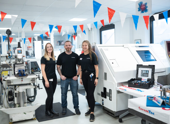 SCHULEWIRTSCHAFT Preisträger CeramTec - drei Mitarbeiter stehen in einem Präsentationsraum zwischen Maschinen der Firma CeramTec