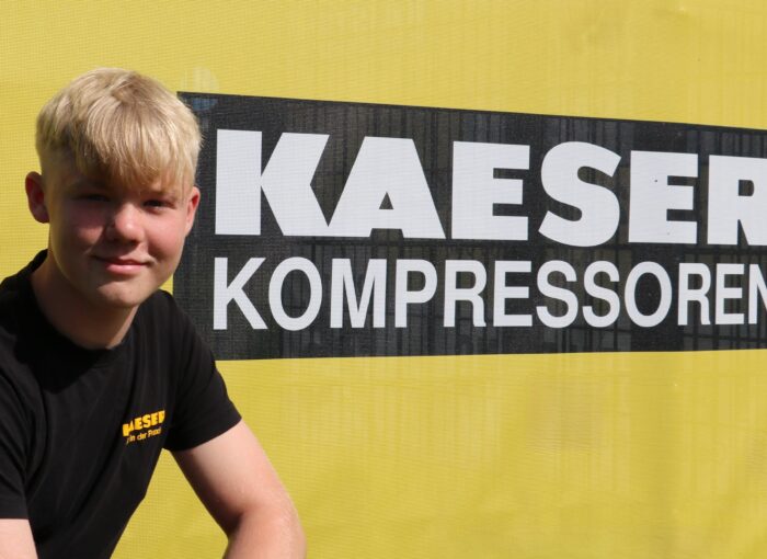 SCHULEWIRTSCHAFT Preisträger KAESER - junger Mann sitzt vor Wand mit Logo von KAESER Kompressoren